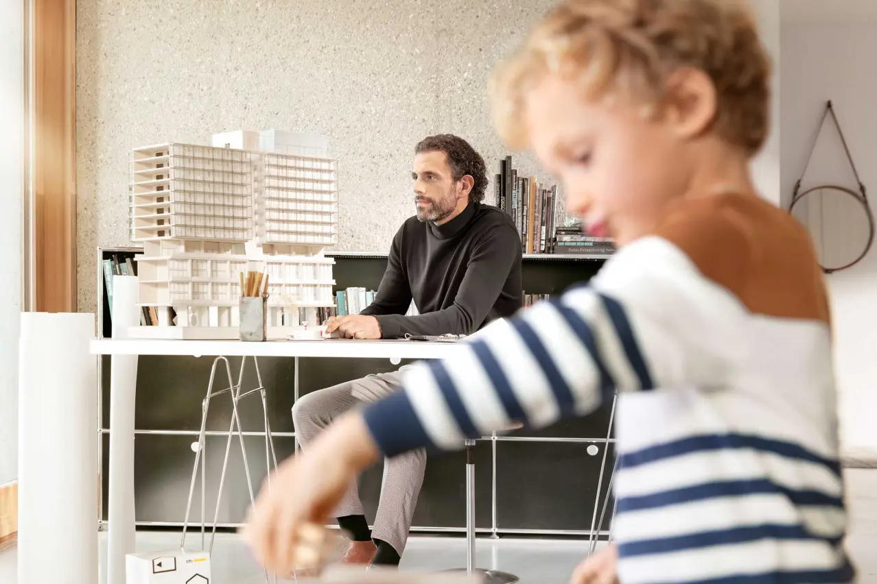 Photo: Fuzzy image of child playing in foreground, father in background, architect sitting at a desk.
